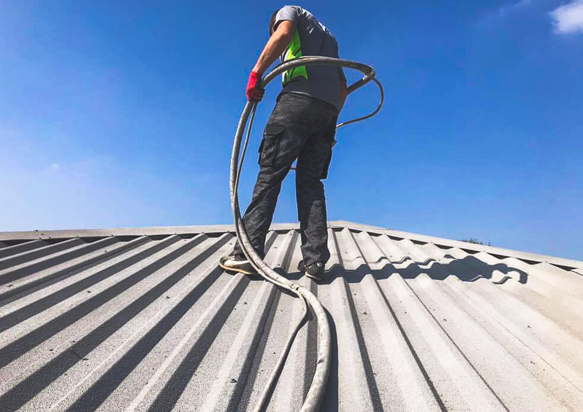 Applicator holding spray equipment on a profiled roof beneath a blue sky
