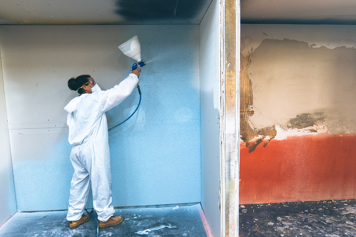 Trainee wearing protective coveralls and a mask applying spray cork to a training wall