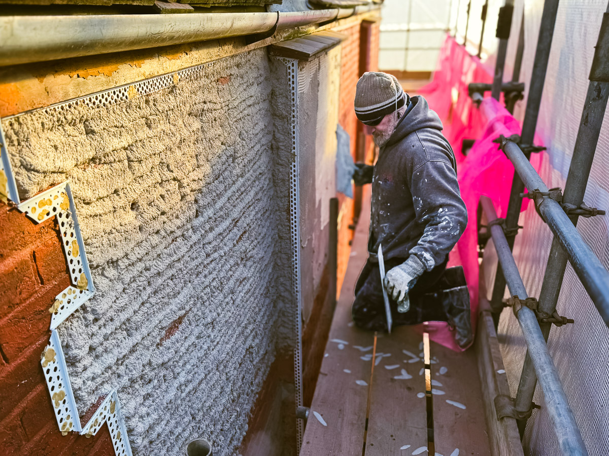 Applicator working on an exterior wall during retrofit, with corner beads and scaffolding visible