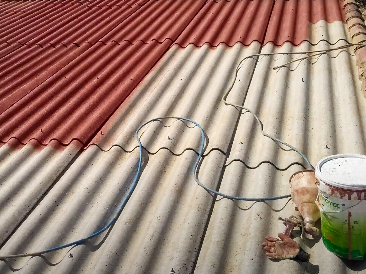 Corrugated roof partly coated in red, with spray equipment and a hose on the uncoated section