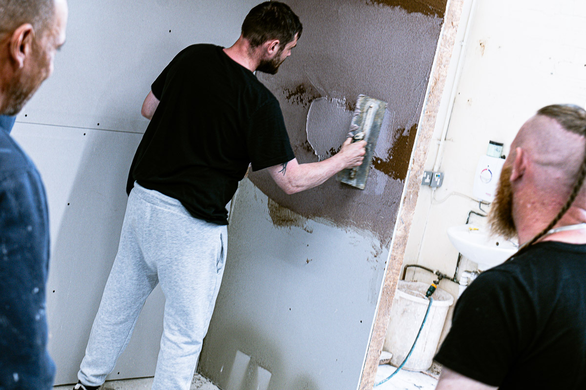 A trainee applies material with a trowel while two people observe